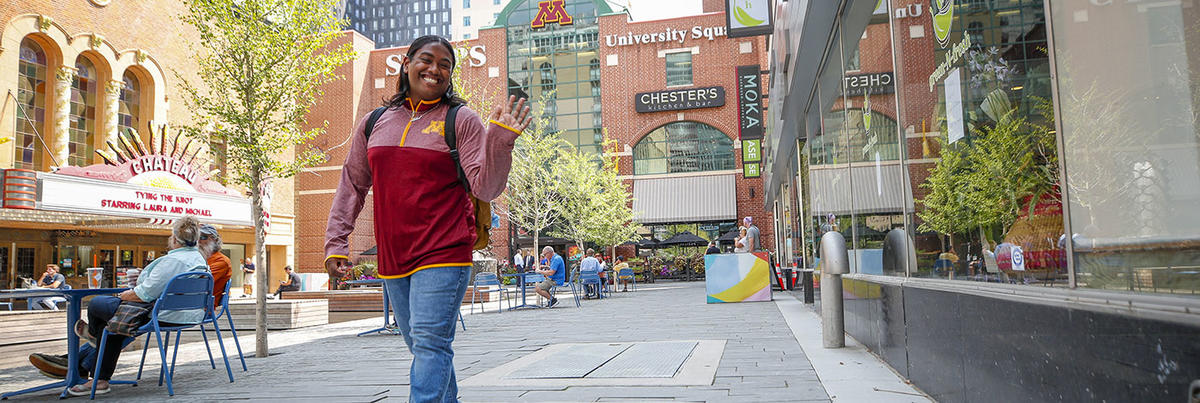 A black student wearing a University of Minnesota sweatshirt is walking in University Square in Rochester and is smiling and waving to someone