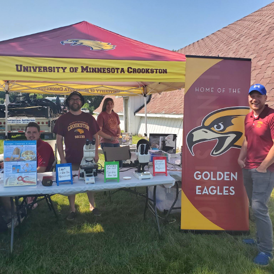 A profile view of a U of M Crookston booth at a summer fair featuring a Golden Eagles team sign