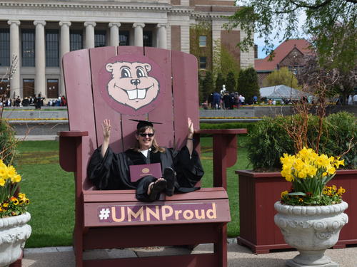 Student in a cap and gown on a chair with Goldy Gopher