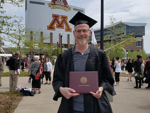 Person posing in cap and gown holding diploma from the University of Minnesota