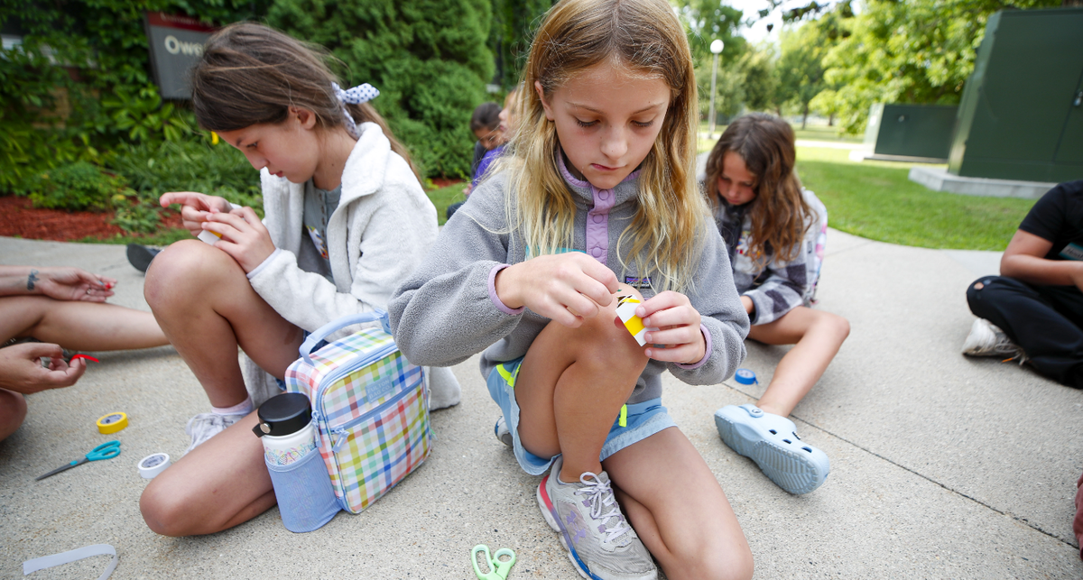 Children sit on a sidewalk doing a science project with paper, tape and scissors