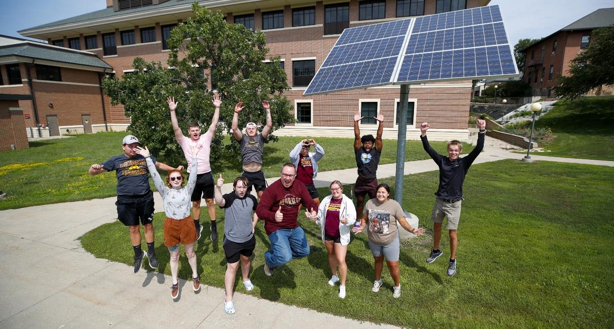 10 students and a teacher leap in the air for a photo in front of a Morris campus building next to a large solar panel 